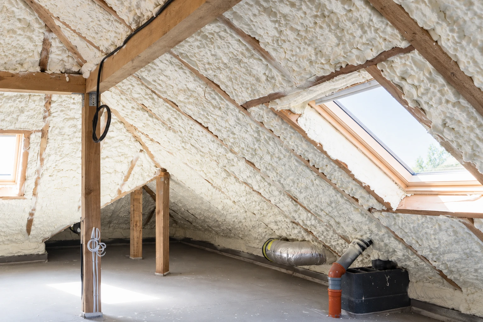 Attic rafters and roof slopes are coated with white spray foam around a skylight and ventilation ducts.