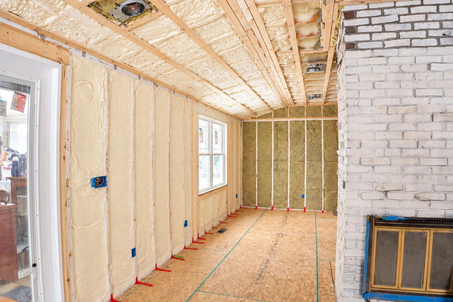 Room under construction with pale spray foam on the walls and ceiling, fiberglass batts in the far wall, and an exposed brick fireplace.