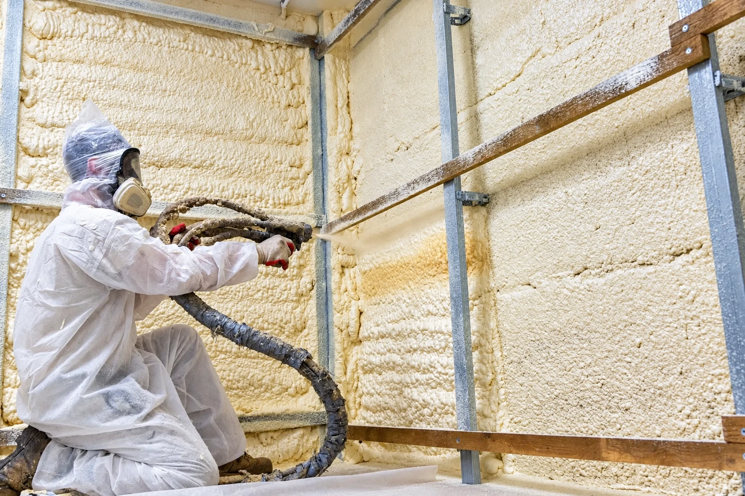 Worker in protective gear sprays closed-cell foam onto a framed wall, coating the spaces between metal supports.