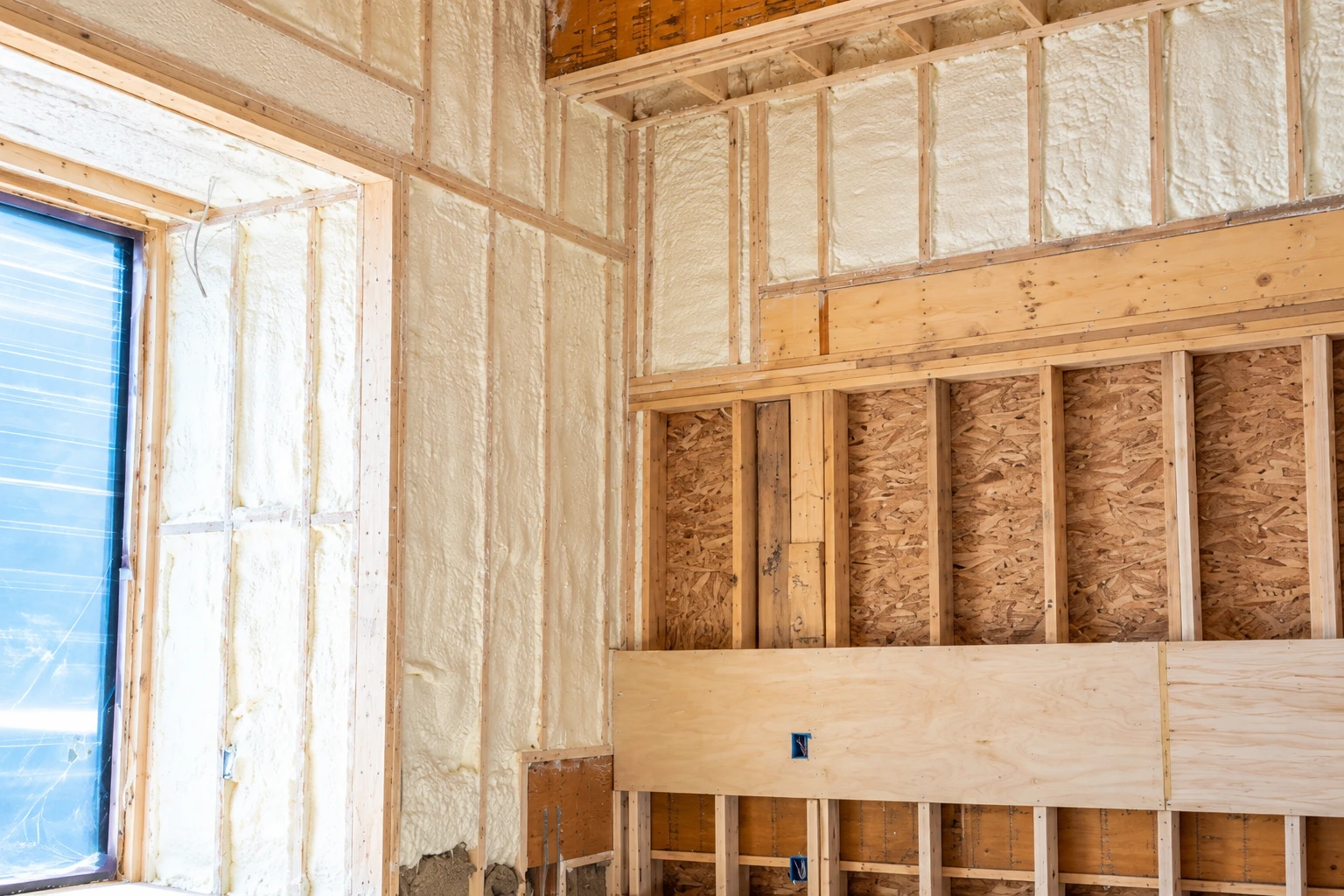 White spray foam fills wall cavities between exposed wooden studs in an unfinished room.