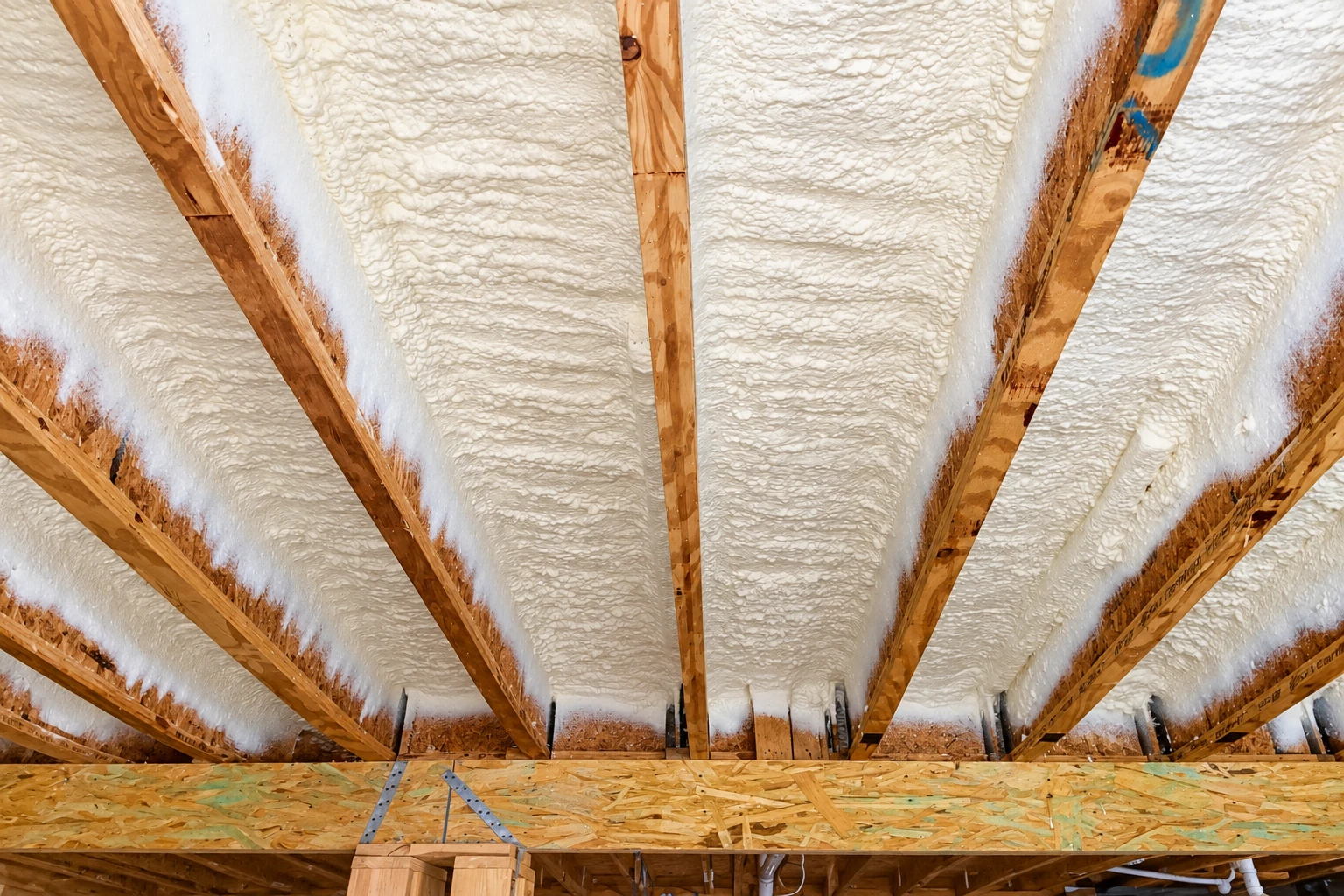 White spray foam fills the spaces between exposed wooden rafters beneath a roof.