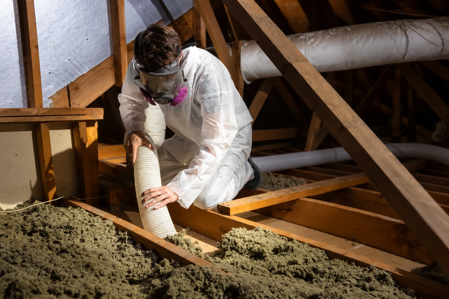 A worker in protective coveralls and a respirator vacuums loose attic insulation with a flexible hose.