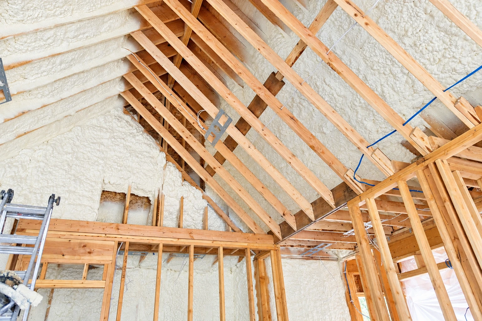 Open-cell foam insulation fills the roof and wall cavities of a house under construction, between exposed wooden framing.