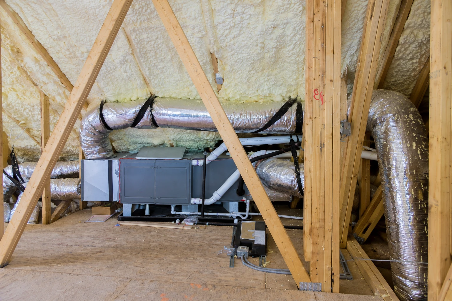 Foam insulation coats the attic roof around exposed wooden trusses, ductwork, and a central HVAC unit.