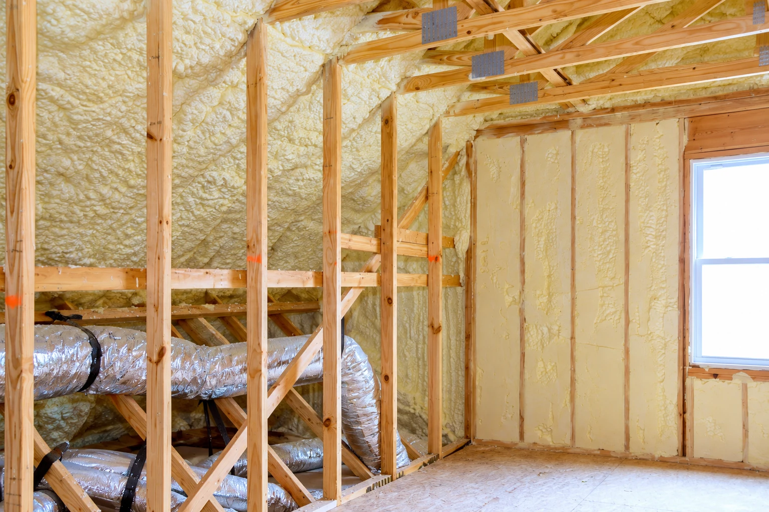Retrofit insulation in an unfinished attic, with yellow spray foam between roof framing and exposed silver HVAC ducts.