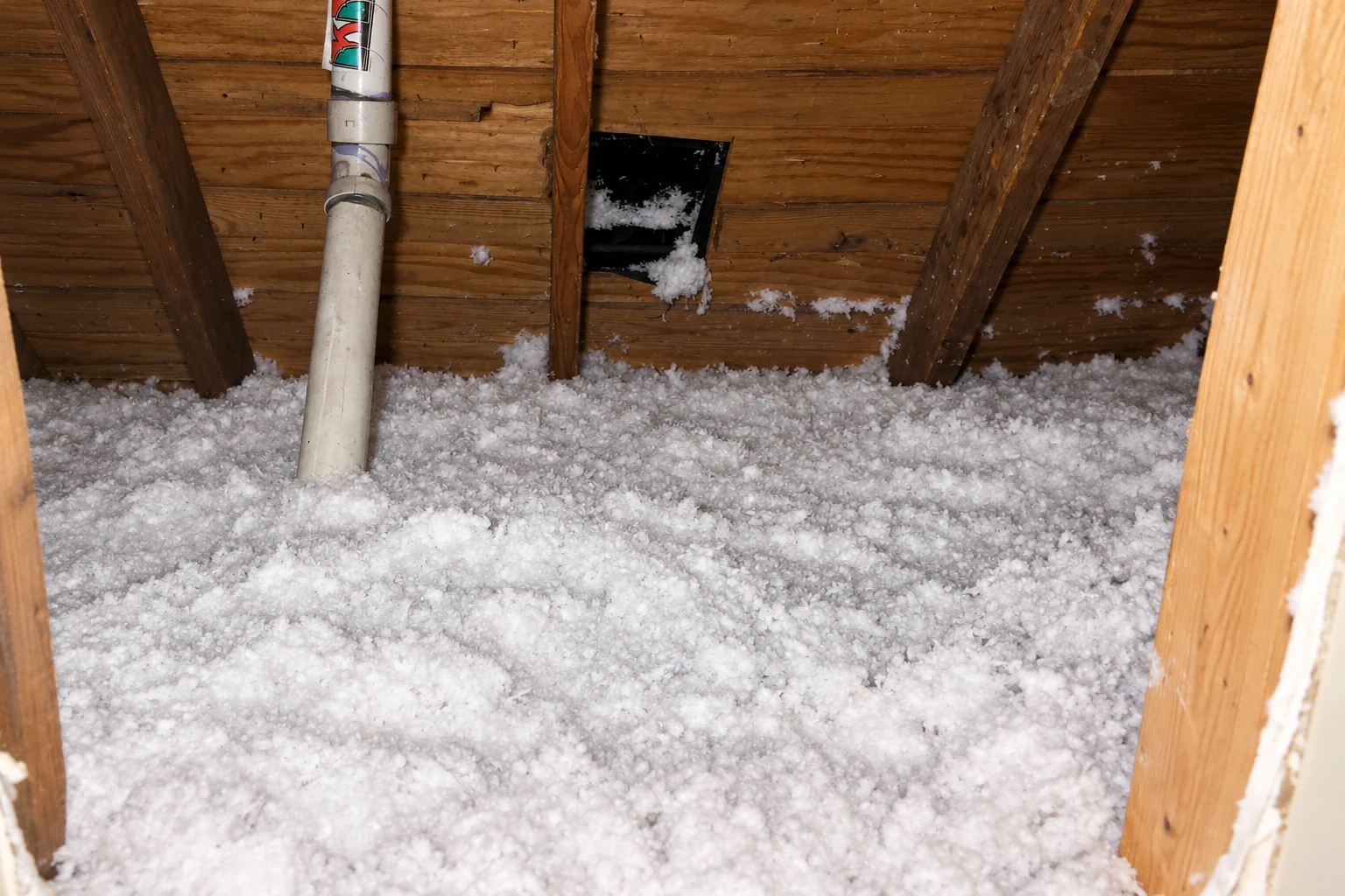 Loose white blown-in insulation covers an attic floor beneath exposed wooden rafters and a vertical pipe.