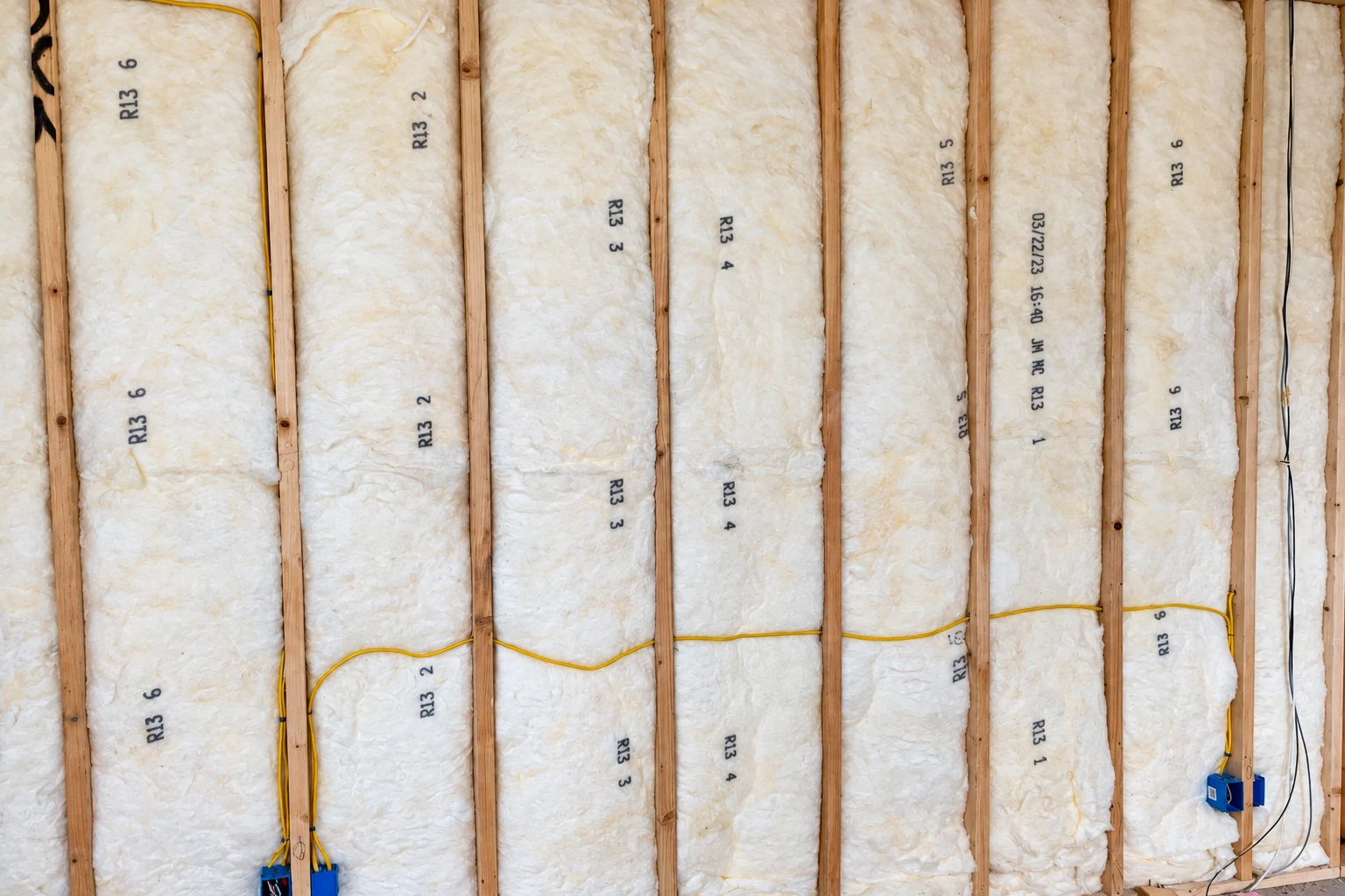 Fiberglass batts fill the bays between exposed wooden wall studs, with yellow wiring running across them.
